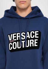 Felpa con cappuccio blu navy realizzata in misto cotone, caratterizzata da un logo stampato audace in bianco e nero con accenti rosa. I lacci del cappuccio sono blu navy.
