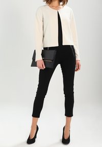Cremefarbene Texturjacke über einem schwarzen Top, kombiniert mit schwarzen, cropped Hosen und schwarzen High Heels. Hält eine schwarze Clutch mit einem goldenen Akzent.