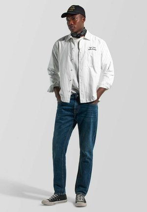 Junger Mann, der ein weißes Nadelstreifenhemd über einem weißen T-Shirt trägt, blaue Jeans, schwarze Mütze, schwarze Turnschuhe und einen schwarzen Halstuch, steht mit den Händen in den Taschen.