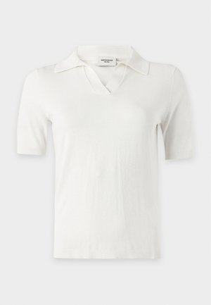 Femme aux longs cheveux noirs portant un polo blanc à manches courtes, une jupe large bleu marine et des ballerines noires pointues avec bride arrière.