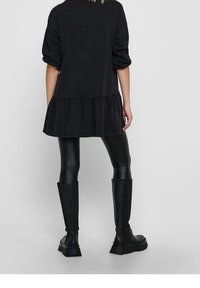 Kobieta ubrana w luźną czarną sukienkę, czarne skórzane legginsy i wysokie czarne botki, widoczna od tyłu na jednolitym tle.