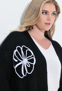 Černý pletený kardigan s velkou bílou květinovou výšivkou na levé straně, pod kterým je hladký bílý top.