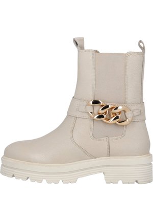 Bottine beige avec une tige en cuir texturé, un talon épais et un accent en chaîne dorée sur la sangle. Elle présente des panneaux élastiques sur les côtés pour un ajustement optimal.