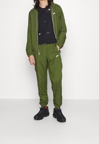 Chándal de nailon verde con chaqueta con cremallera y pantalones de cintura elástica. Camiseta gráfica negra con logos. Zapatillas negras con detalles rojos.