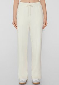 Crème-kleurige sweatpants met een trekkoord in de taille, brede pijpen en subtiele verticale naden, gecombineerd met witte sneakers en een neutrale achtergrond.