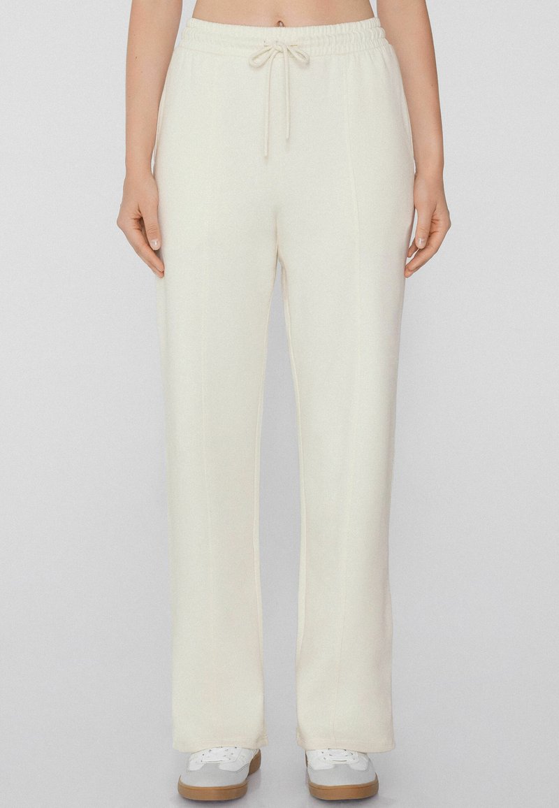 Crème-kleurige sweatpants met een trekkoord in de taille, brede pijpen en subtiele verticale naden, gecombineerd met witte sneakers en een neutrale achtergrond.