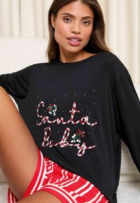 Fekete, hosszú ujjú póló "Santa Baby" felirattal piros és fehér cukornádró betűkkel, zöld magyallevelekkel és fehér pöttyökkel díszítve. Pár piros csíkos rövidnadrág.