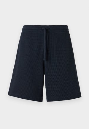 Homme aux cheveux blonds courts portant une chemise à manches courtes à motifs bleu, vert et beige, un short bleu marine et des sandales marron, debout les mains dans les poches.