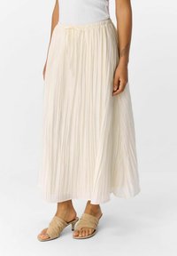 Falda maxi plisada en beige claro con cintura elástica y cordón. La tela tiene una textura suave y cae con delicadeza.