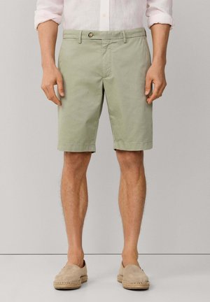 Hombre con pantalones cortos hasta la rodilla de color verde claro, camisa blanca de manga larga remangada y zapatos beige sin cordones, de pie contra un fondo liso.