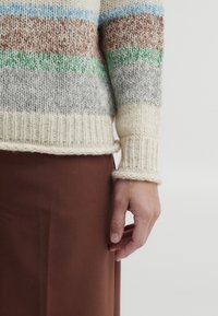 Hand rustend langs het lichaam, gekleed in een gestreepte gebreide trui met kleuren crème, blauw, bruin, groen en grijs, en een bruine rok of broek.