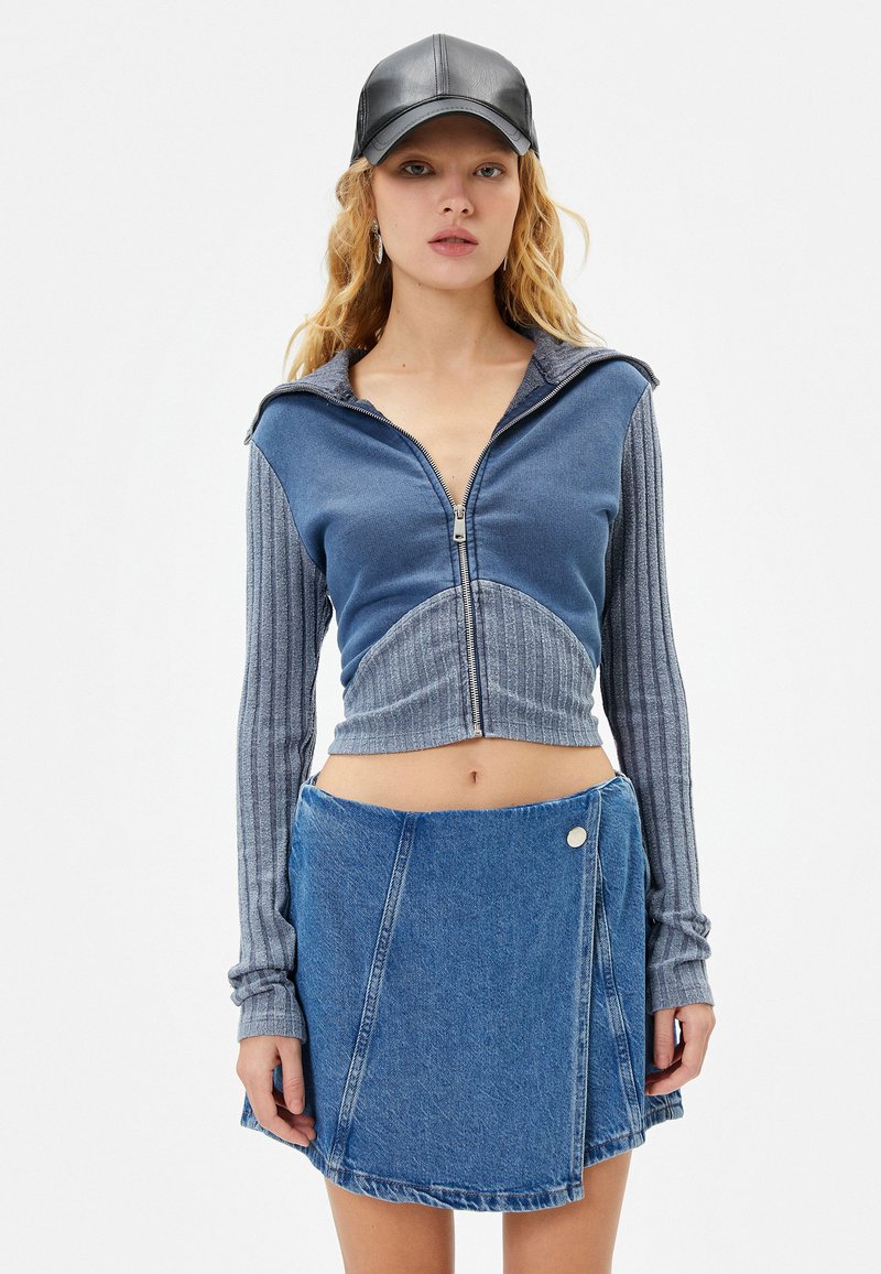 Blaues, kurz geschnittenes Zip-Hoodie mit gerippten Ärmeln, kombiniert mit einem Jeans-Wickelrock mit Druckknopfverschluss und einem fransigen Saum. Schwarze Mütze auf dem Kopf.