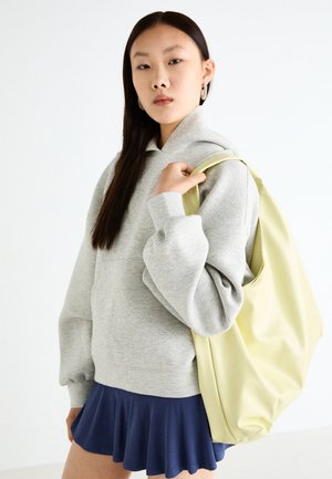 Sweat-shirt oversized gris avec une poche kangourou, associé à une jupe évasée marine. Accessoire : grand sac à dos jaune clair, texture lisse.