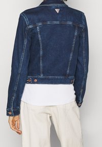 Denim jacket in dark blue with a structured design, copper buttons, and a triangular logo at the back. Features long sleeves and a cropped cut.