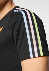 T-shirt noir à coupe ajustée, tissu texturé, avec des rayures colorées en vert, rose, bleu et noir le long des épaules.