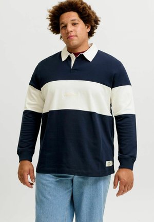 Giovane uomo con capelli ricci che indossa una maglia da rugby a maniche lunghe blu navy e bianca e jeans azzurri, in piedi davanti a uno sfondo semplice.