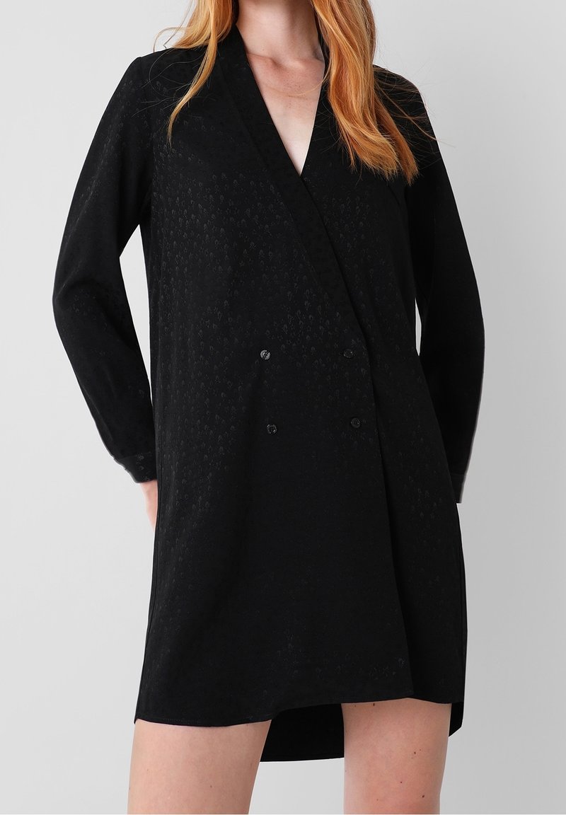 Femme portant une robe blazer noire texturée à manches longues avec des boutons croisés, debout devant un fond gris uni.