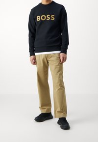 Hombre vistiendo un sudadera negra con el texto "BOSS", pantalones beige, zapatillas negras y camiseta blanca debajo, contra un fondo blanco liso.