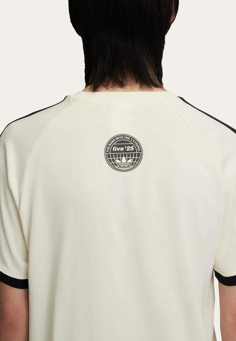 Camicia color crema con dettagli neri sulle maniche. Presenta un logo circolare sul retro con testo e un elemento grafico. Tessuto leggero.