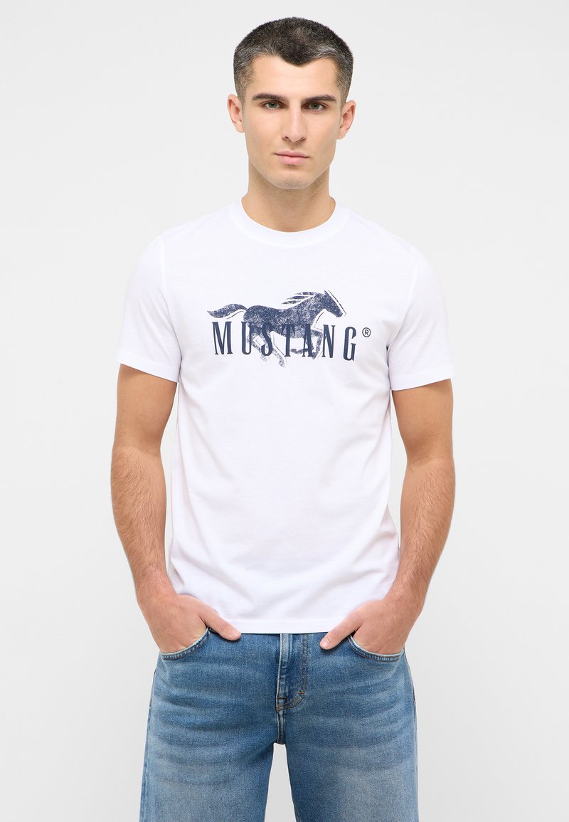 T-shirt en coton blanc avec un graphique de cheval en marine et le texte "MUSTANG", à manches courtes et col rond, associé à un jean bleu clair.
