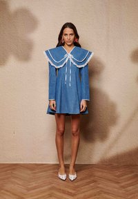 Τζιν φόρεμα με μεγάλο άσπρο γιακά διακοσμημένο με δαντέλα και κλείσιμο με κορδόνι, με μακριά μανίκια και φλοράλ φούστα. Φορεμένο με παπούτσια με σχέδια.