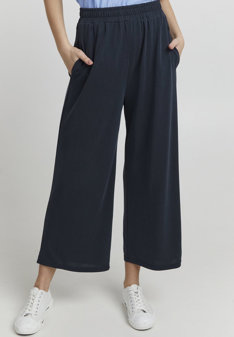 Pantaloni a gamba larga blu navy realizzati in tessuto morbido; presentano un elastico in vita e tasche laterali, abbinati a sneakers bianche.