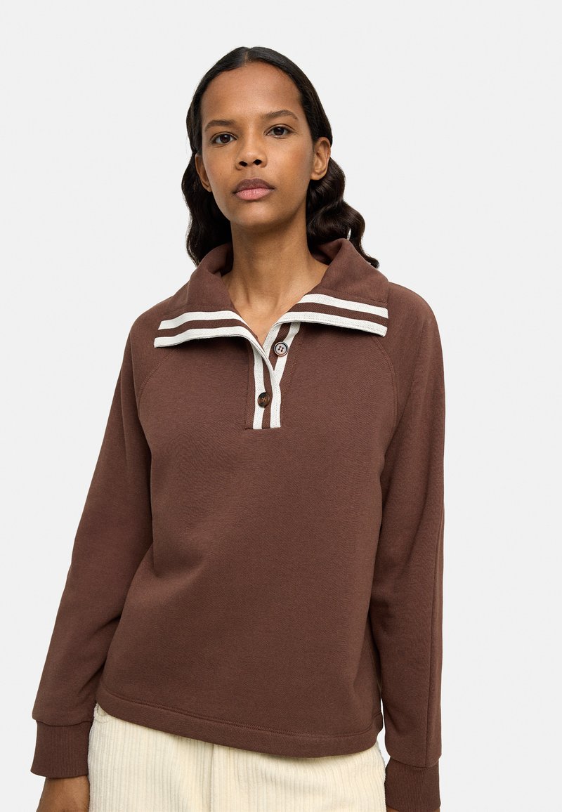 Brązowy sweter z kołnierzem w białe paski, zapinany na dwa guziki, z długimi rękawami, wykonany z teksturowanej tkaniny. Stylizowany z jasnymi spodniami.
