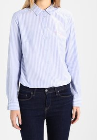 Frau, die ein hellblaues und weißes, senkrecht gestreiftes Hemd mit Knöpfen trägt, in dunkle blaue Jeans gesteckt, die sichtbare Vorderentaschen haben.