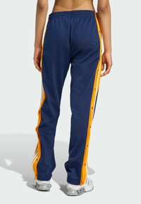 Navy blue sweatpants with orange side stripes, snap buttons, and an elastic waistband. Smooth, lightweight material with a relaxed fit.