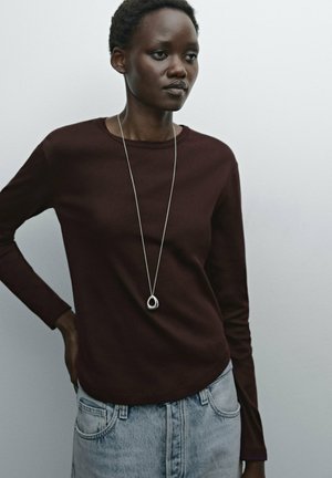 Bruine langermige top in een aansluitend ontwerp, gecombineerd met lichtblauwe high-waisted denim jeans. Het model draagt een zilveren hangerketting.