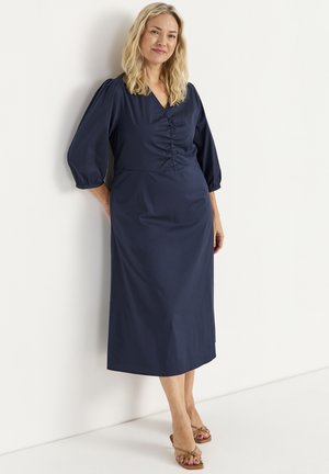 Vrouw met blond haar, gekleed in een marineblauwe midi-jurk met V-hals en plooien, en beige sandalen met hak, staand tegen een witte muur.