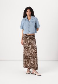 Jaqueta de ganga azul cortada com mangas curtas, combinada com uma saia longa de padrão de leopardo e sapatos slip-on de cor clara.
