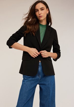 Blazer negro de tejido suave, con un diseño ajustado, dos bolsillos frontales y cierre de un solo botón. Lleva puesto un top verde.