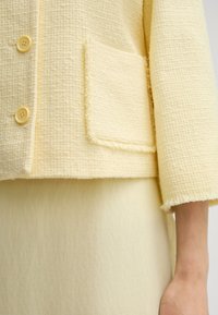 Giacca gialla con texture e bordi sfilacciati, due bottoni e una tasca con pattina. Indossata sopra una gonna gialla chiara dalla finitura liscia.