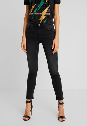 Femme portant un jean slim noir, un t-shirt graphique noir avec un motif éclair et des sandales noires à talons hauts avec brides à la cheville.