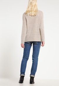 Beige Strickpullover mit langenÄrmeln, lockerer Passform und abgerundetem Saum, kombiniert mit schmalen, dunkelblauen Jeans und schwarzen Stiefeletten.