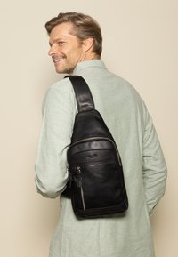 Schwarze Lederschultertasche mit glatter Oberfläche, ausgestattet mit mehreren Reißverschlusstaschen und einem breiten, verstellbaren Riemen, der über der Schulter getragen wird.