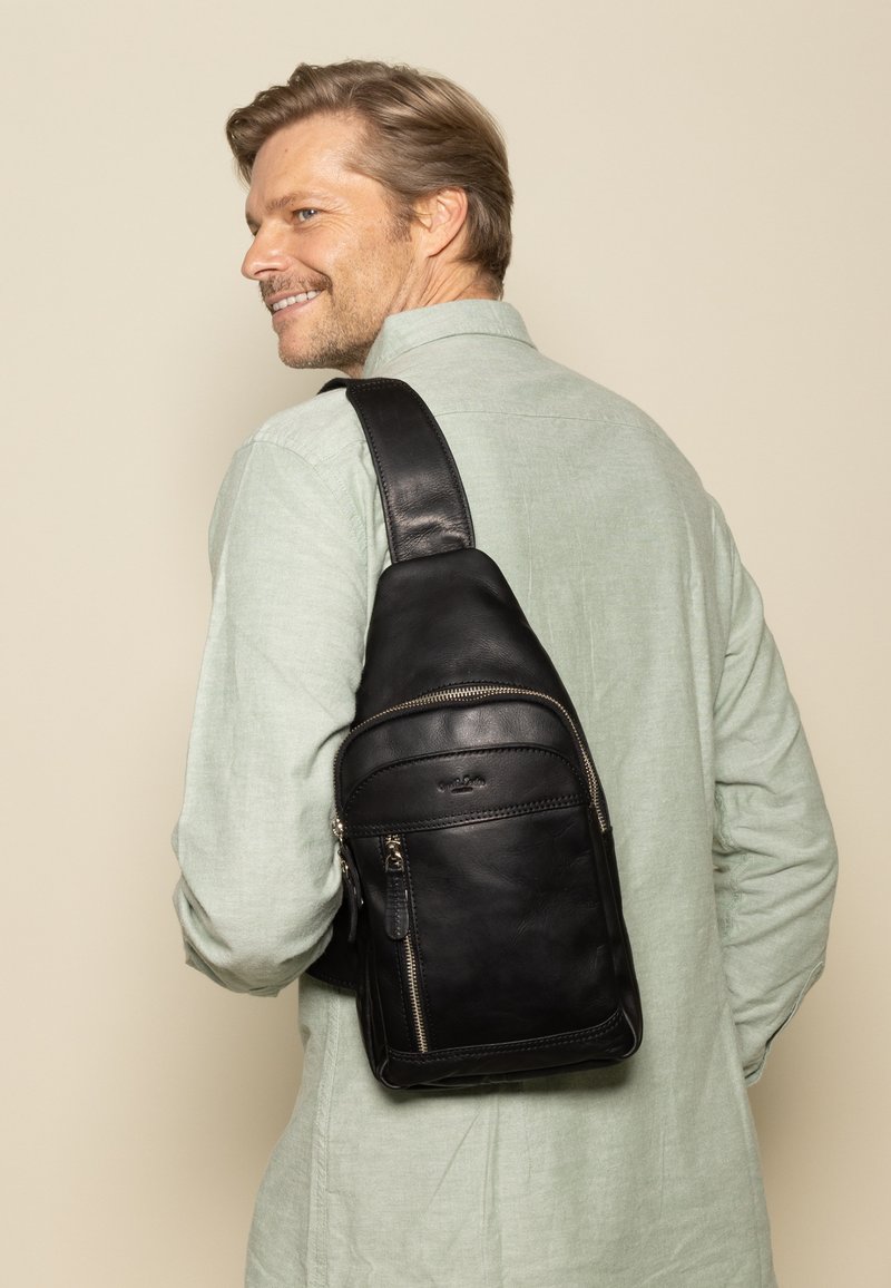 Schwarze Lederschultertasche mit glatter Oberfläche, ausgestattet mit mehreren Reißverschlusstaschen und einem breiten, verstellbaren Riemen, der über der Schulter getragen wird.