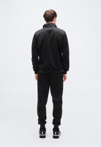 Μαύρο αθλητικό σετ με φούτερ με φερμουάρ και παντελόνια με ελαστική μέση. Διαθέτει λεία, ματ υφή και εφαρμοστά μανσέτες στα μανίκια και στους αστραγάλους.