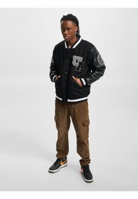 Veste varsity noire avec des manches en cuir, lettres grises brodées "UA", poignets rayés ; associée à un pantalon cargo en velours côtelé marron et des baskets noires.