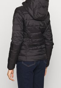 Svart puffjacka med huva, med ett quiltat mönster och en slät, glänsande yta. Passformen är figursydd vid midjan, med elastiska ärmslut.