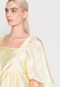 Vestido amarelo claro com decote quadrado e mangas bufantes, feito de um tecido brilhante e texturizado. Apresenta detalhes franzidos na cintura.