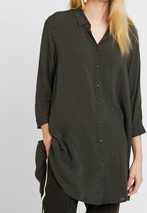 Woman wearing a long black button-up shirt with white polka dots, three-quarter sleeves, and black pants with white side stripes.