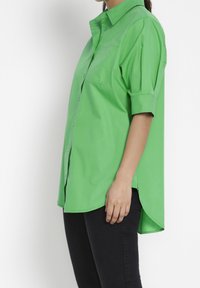 Grön skjorta med knappar i en slät tyg, med korta ärmar, krage och en enskild bröstficka, med en rundad fåll.