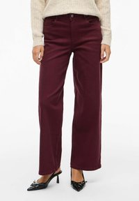 Jeans a gamba larga bordeaux con chiusura frontale a bottoni, cinque tasche e una texture di cotone liscia. Abbinati a décolleté nere a punta.