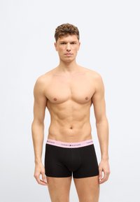 Άνδρας χωρίς μπλούζα με κατσαρά μαλλιά, που φοράει μαύρα boxer briefs με ροζ ζώνη μέσης Tommy Hilfiger, στέκεται μπροστά σε ένα απλό λευκό φόντο.