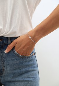 Silberne Armbanduhr mit einem kleinen Sternanhänger, filigrane Kette, am Handgelenk getragen, kombiniert mit einem weißen Hemd und blauen Jeans.