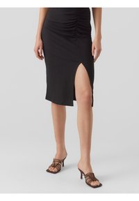 Jupe noire ajustée avec détail froncé, longueur genoux, dotée d'une fente sur le côté. Associée à des mules à talons strappés marron. Tissu lisse et texturé.