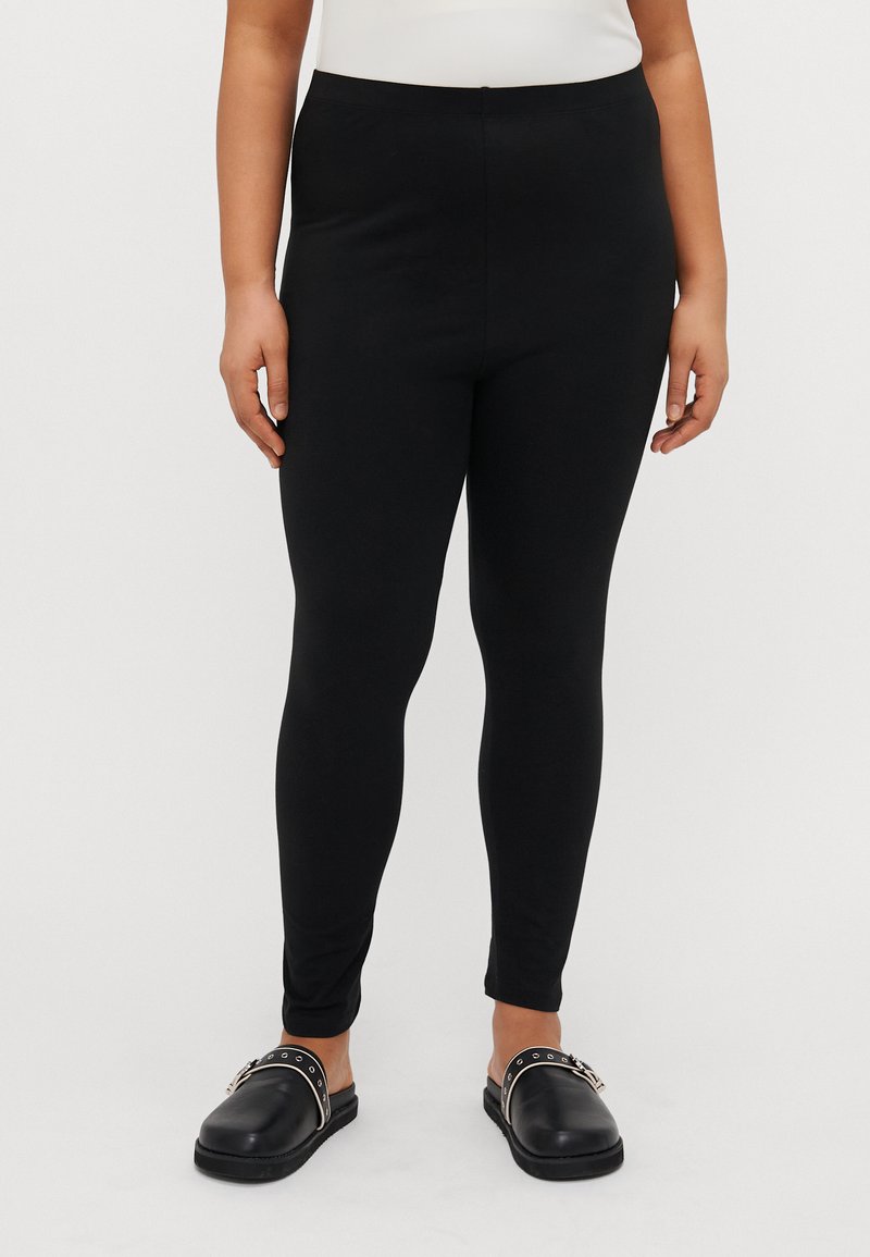 Zwarte leggings van een gladde, elastische stof, met een elastische tailleband en een aansluitende pasvorm, gecombineerd met zwarte instap schoenen.
