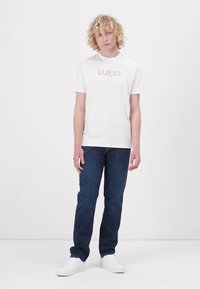 Fehér pamut póló "GUESS" logóval világos színekben, sötétkék farmernadrággal és fehér sportcipővel párosítva. A modell egy sima háttér előtt áll.